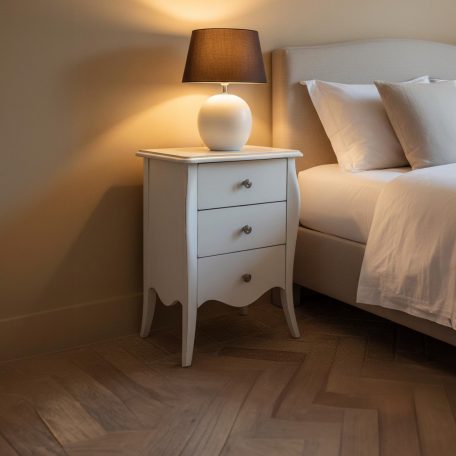 PR-4253 Ein weißer Nachttisch mit drei Schubladen und einer Lampe neben einem Bett.